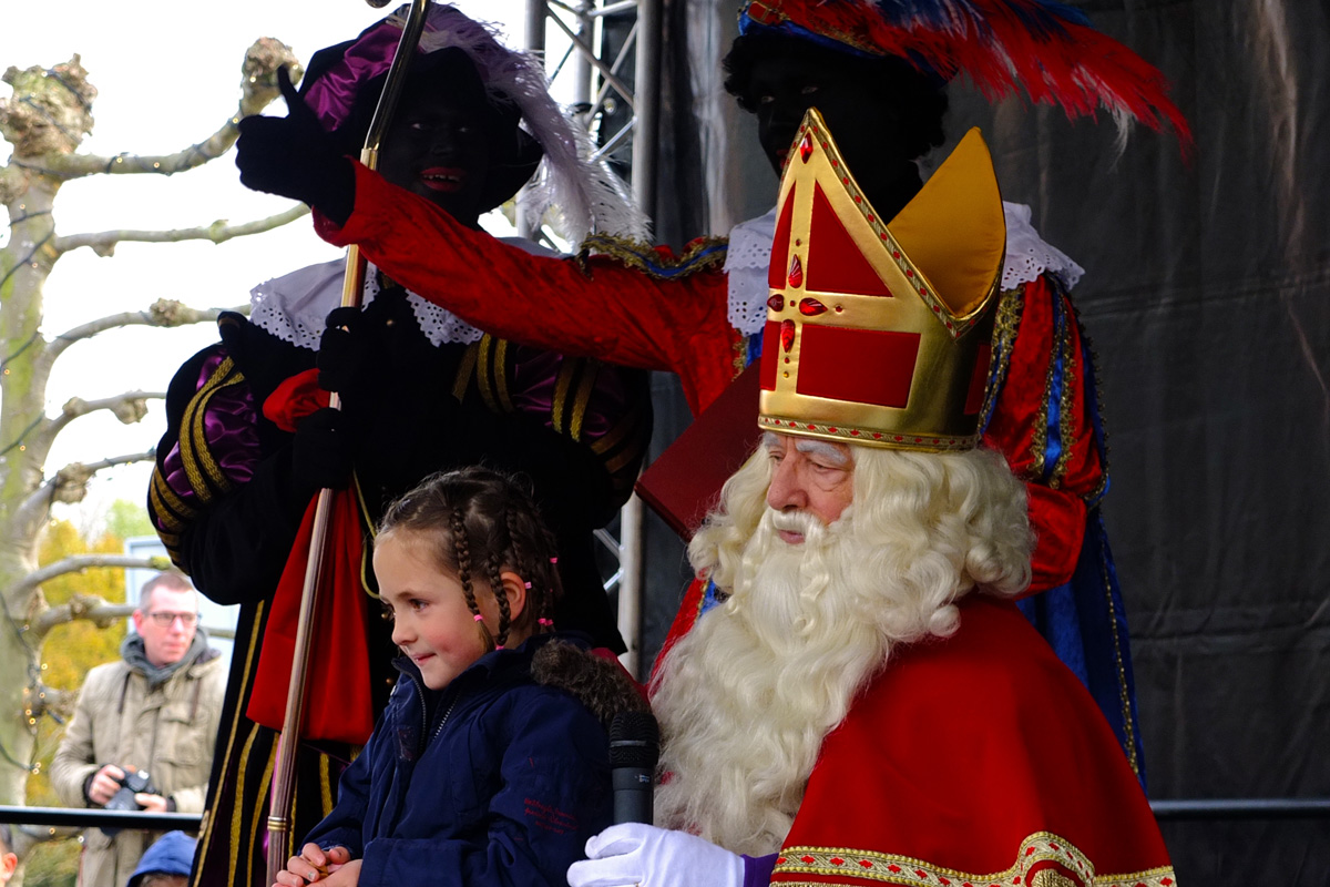 Sinterklaas con una niña sentada en su regazo, acompañado por Pieten durante una celebración en los Países Bajos.