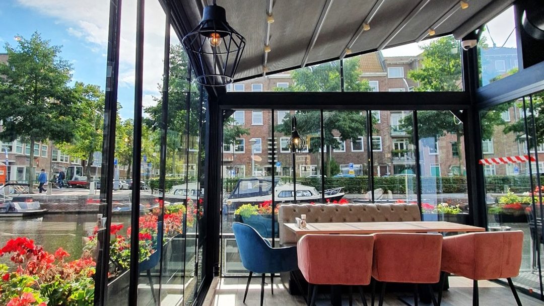 Zona acristalada del Florya Restaurant & Café en Ámsterdam con vistas al canal y decoración acogedora.