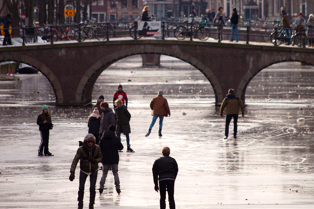 Personas patinando sobre un canal helado en Ámsterdam, con un puente de ladrillo cruzando el canal en un día de invierno.