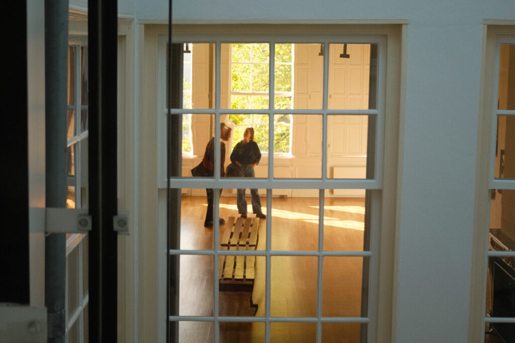 Vista desde una ventana del Huis Marseille, con dos visitantes observando una sala del museo iluminada por el sol de invierno.