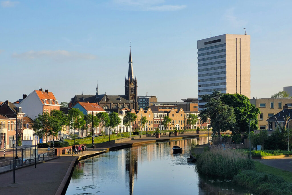 Canal de Delft al atardecer con edificios históricos frente al agua y la torre de la Nieuwe Kerk al fondo, junto a un edificio moderno visible desde el canal.