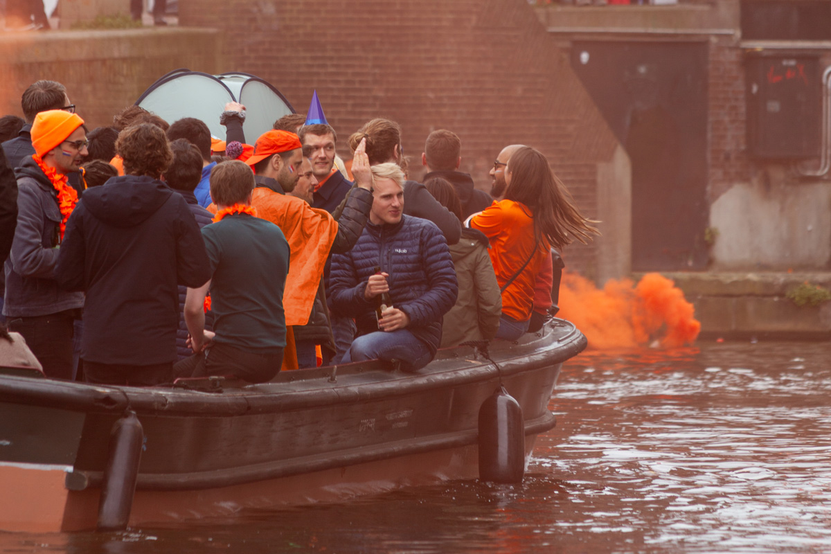 Jóvenes vestidos de naranja celebrando el Día del Rey en un barco en los canales de Ámsterdam, con humo naranja alrededor y ambiente festivo.