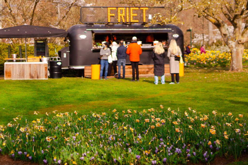 Tulipanes en primer plano con un puesto de comida al fondo dentro del parque Keukenhof; escena típica de una pausa gastronómica durante la visita.
