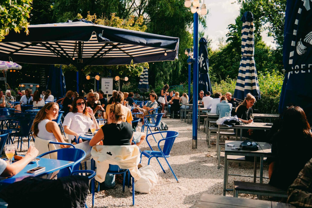 Terraza del Proeflokaal ’t Blauwe Theehuis en Vondelpark con ambiente primaveral, mesas ocupadas y vegetación alrededor.
