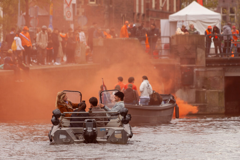 Barcos con jóvenes vestidos de naranja celebrando el Día del Rey en los canales de Ámsterdam, con humo naranja en el aire y ambiente festivo.