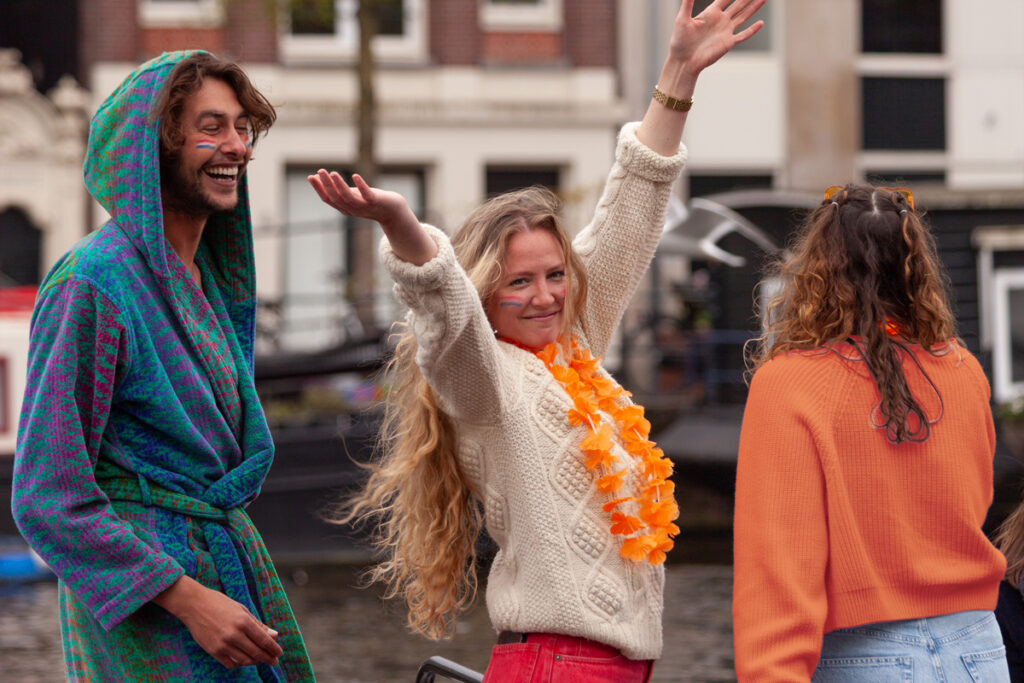 Dos chicas y un chico celebran el Día del Rey en un barco en los canales de Ámsterdam, vestidos de naranja y con accesorios festivos.