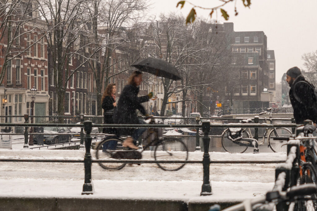 Mujer en bicicleta cruzando un puente en Ámsterdam cubierto de nieve, con la ciudad al fondo en invierno.