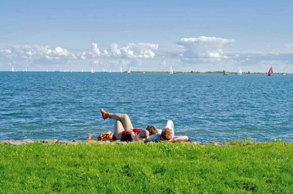 Pareja tumbada en el césped en Volendam observando barcos de vela en el mar en un día de verano.