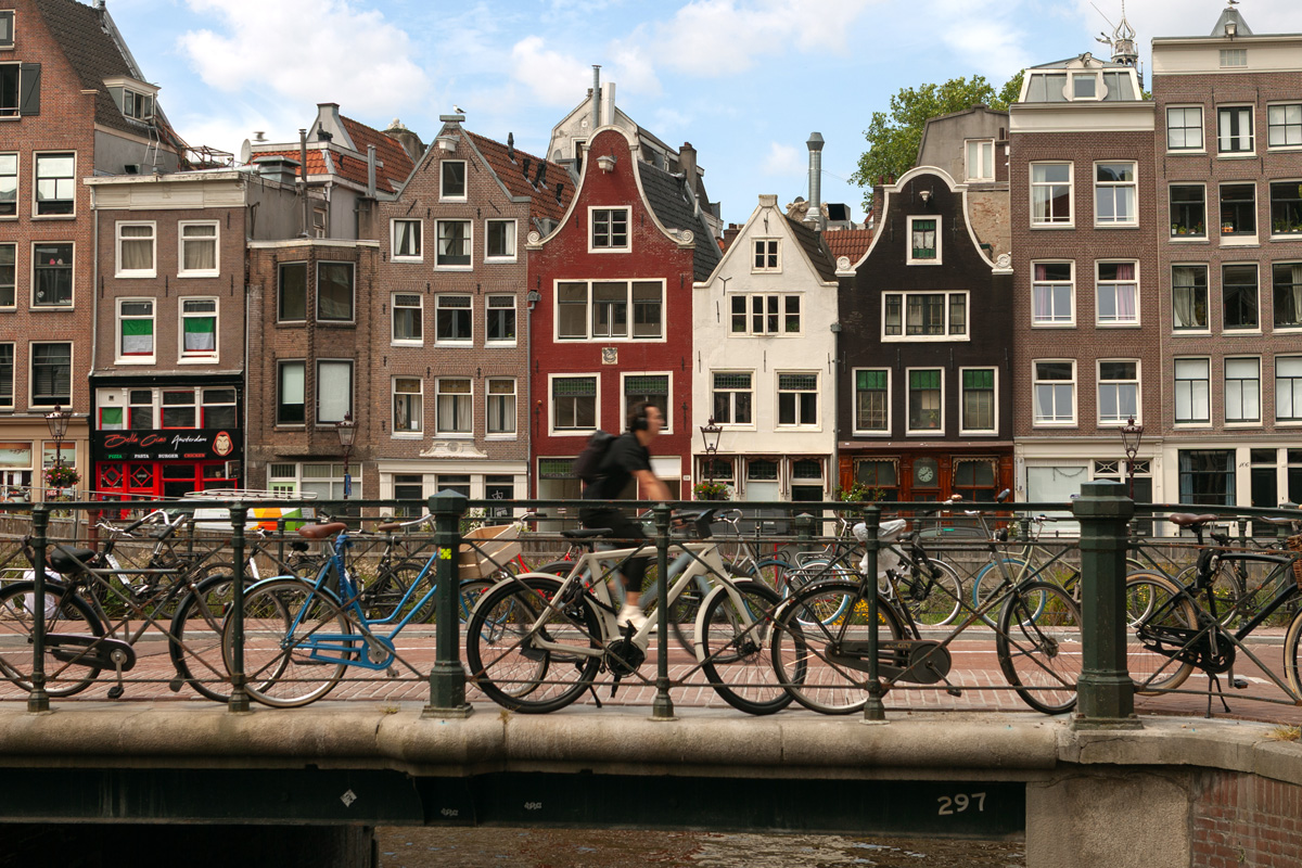 Puente de canal en Ámsterdam con bicicletas aparcadas y un ciclista pasando, con edificios de colores al fondo en un día soleado.