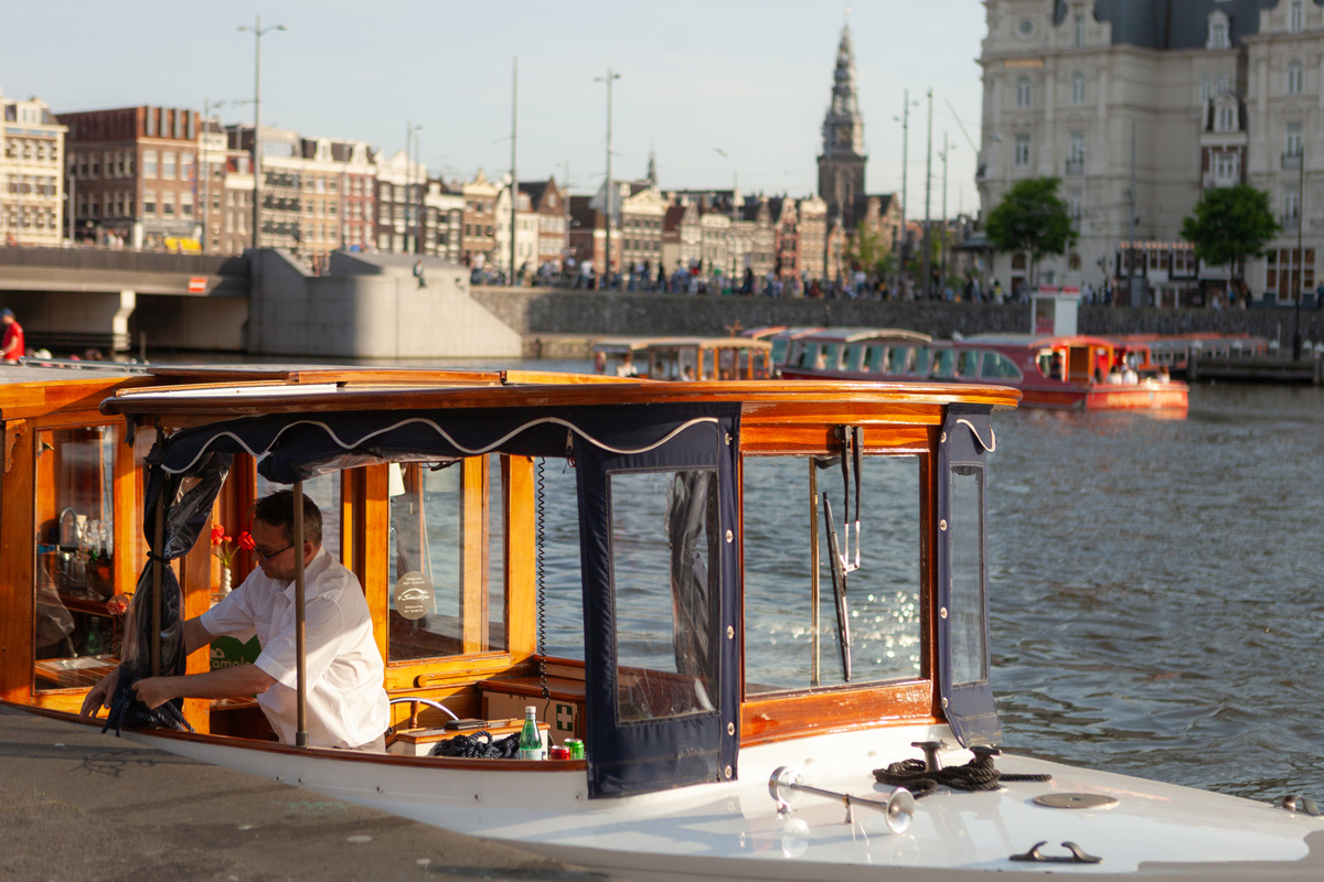 Barco de Camaleón Tours navegando por los canales de Ámsterdam con el patrón a bordo y el centro histórico al fondo.