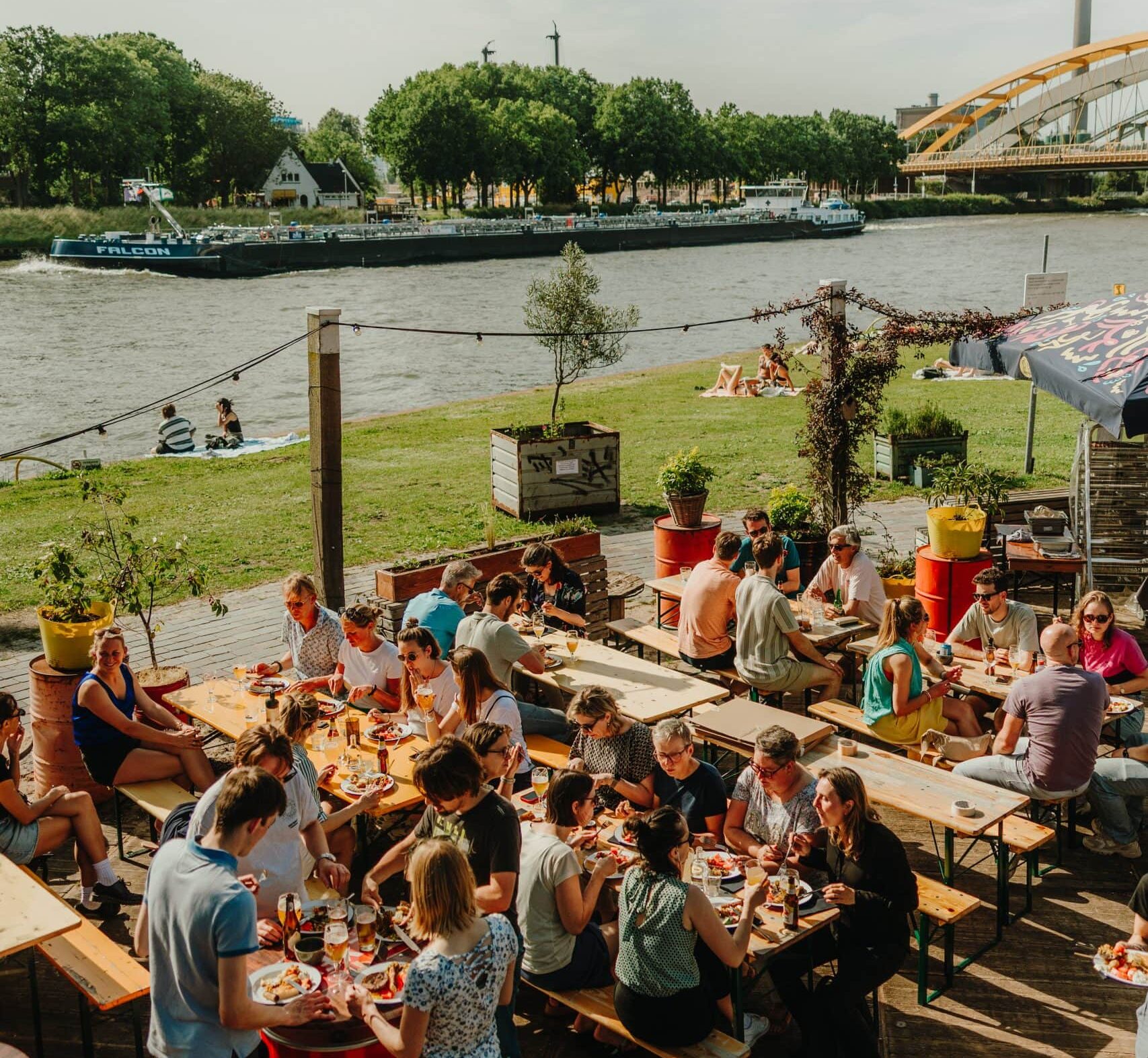 Terraza de SOIA en Utrecht con personas disfrutando del sol en bancos junto al canal y gente descansando en el césped.