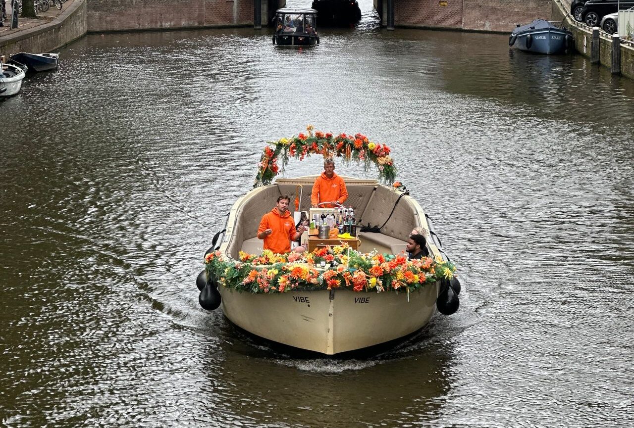 Barco decorado con flores navegando por un canal de Ámsterdam con dos personas vestidas de naranja a bordo.