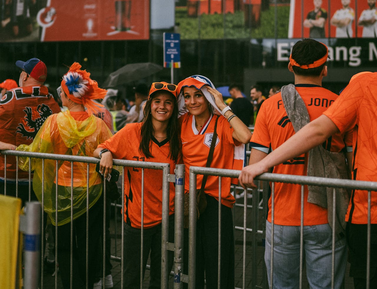 Dos aficionadas vestidas de naranja frente a un estadio de fútbol en los Países Bajos, rodeadas de seguidores neerlandeses.
